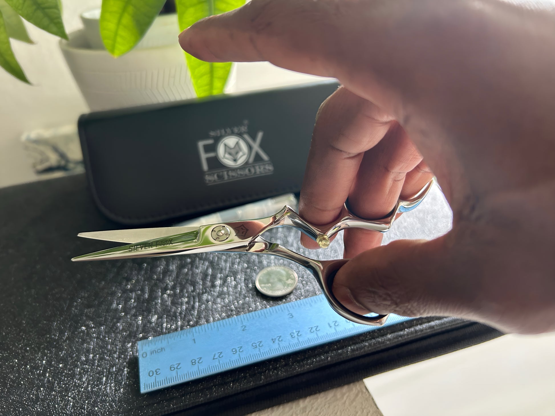 Top Down view of Silver Fox Scissors Model SF28 being held in the right hand, with a ruler, coin, oil canister, and carrying case resting flat on top of a display case.